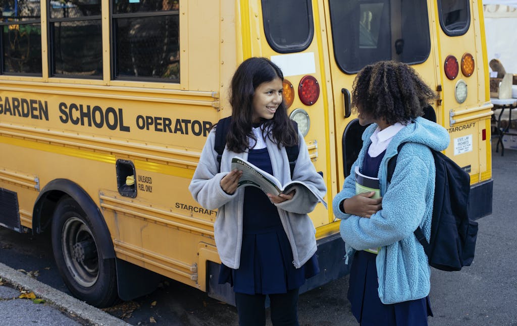 Happy multiracial classmates with backpacks standing near yellow school bus and communicating after school while looking at each other